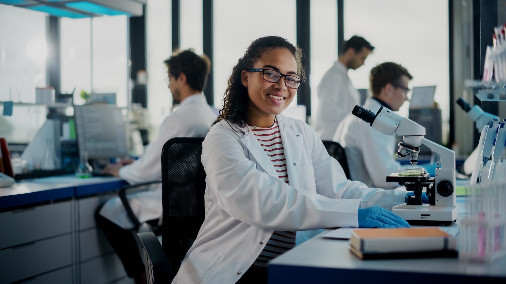 A Mohs technician in a lab coat smiles while analyzing a sample with a microscope in a softly lit lab environment.