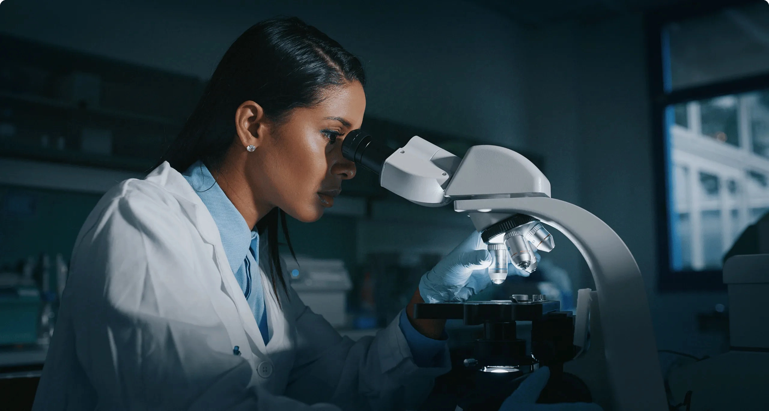 A Mohs technician, in a lab coat, examines a sample using a microscope in a dimly lit laboratory.